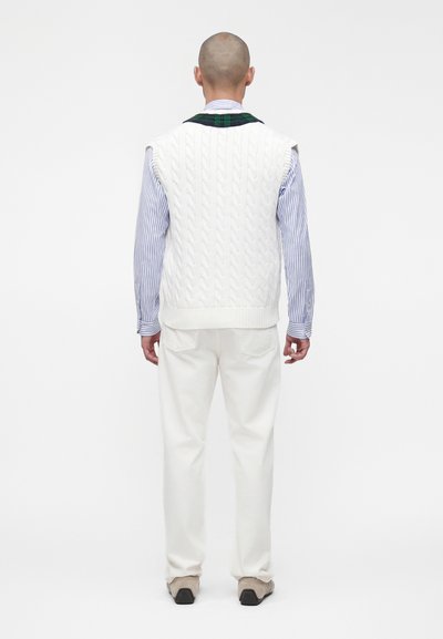 Homme de dos portant un gilet blanc en maille torsadée sur une chemise rayée bleue, un pantalon blanc et des chaussures beiges sur un fond uni.