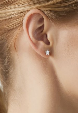 Primer plano de una oreja izquierda con un pequeño pendiente de tachuela plateada en forma de estrella, sobre piel clara y cabello rubio.