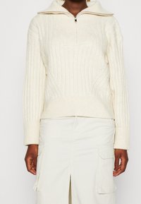 Krémový žebrovaný pletený svetr s polovičním zipem a širokým límcem. Má přiléhavý pas a dlouhé rukávy. Texturovaný, pohodlný design.