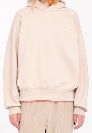 Personne portant un sweat à capuche beige oversize et un pantalon en velours côtelé beige, debout devant un fond blanc.