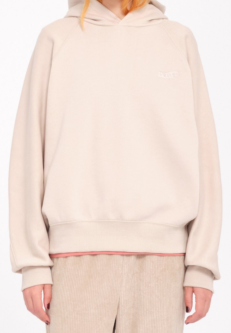 Personne portant un sweat à capuche beige oversize et un pantalon en velours côtelé beige, debout devant un fond blanc.
