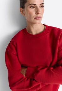 Červená mikina vyrobená z mäkkej, textúrnej tkaniny. Má okrúhly výstrih, spadnuté ramená a dlhé rukávy s rebrovanými manžetami.