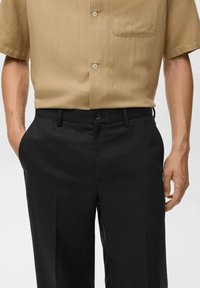 Camicia beige a maniche corte con taschino sul petto, abbinata a pantaloni neri su misura con pieghe frontali e chiusura con bottone.