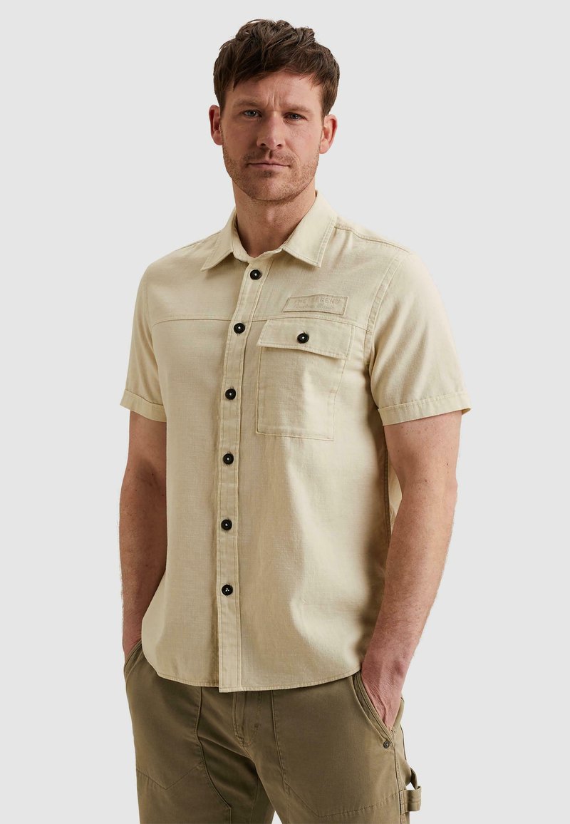 Homme portant une chemise beige à manches courtes avec poche avant et un pantalon vert olive, debout les mains dans les poches devant un fond uni.
