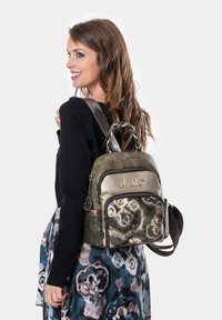Frau mit langen braunen Haaren lächelt, trägt ein schwarzes Oberteil und einen gemusterten Rock und hält einen kleinen metallischen Rucksack mit Blumenmuster.