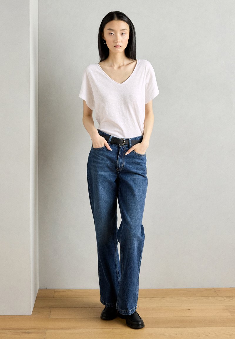 Biała koszulka z dekoltem w serek i luźnymi rękawami, zestawiona z niebieskimi jeansami o wysokim stanem i czarnym obuwiem, pozująca na neutralnym tle.