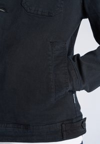 Giacca di jeans nera con mano nella tasca laterale, dettaglio di bottoni visibili all'orlo inferiore e cuciture sulla tasca del petto.