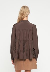 Brown tiered blouse with a collar and puffed long sleeves. Smooth fabric with pleats, featuring buttoned cuffs and a relaxed shape.