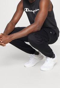 Svart ärmlös topp med "Champion"-logotyp, svarta joggerbyxor med sidofickor och vita sportskor med texturerad detalj.