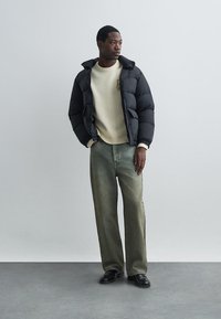 Melnā puffa jaka ar kapuci, bēša džemperis, zaļas plašas bikses un melnas kurpes, stāvot pret neitrālu fona.