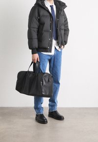 Schwarze Daunenjacke mit Kapuze, weißes Hemd, blaue Jeans, schwarze Lederschuhe und eine große schwarze Reisetasche mit Griffen.