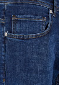 Gros plan d'une poche de jean en denim bleu avec des coutures orange et un rivet métallique visible dans le coin.