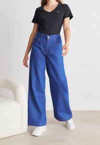 Modré široké džínsy s vysokým pasem a oranžovým šitím, kombinované s černým tričkem s V-výstřihem a nošeny s bílými teniskami.