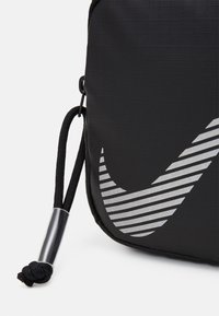 Borsa nera realizzata in tessuto testurizzato, con un grande logo Nike a strisce argentate, cordino nero regolabile e dettagli in metallo.