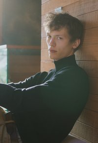 Μαύρο πουλόβερ με ψηλό λαιμό από υφασμάτινη υφή, leaning against a wooden wall, μαλακό φωτισμό που τονίζει τα χαρακτηριστικά του προσώπου και την άνετη στάση.