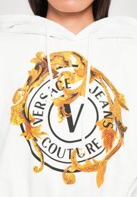 Felpa bianca con cappuccio caratterizzata da un logo circolare con la scritta "Versace Jeans Couture", decorata con elementi grafici dorati e neri e strass.