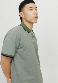 Pilkas polo marškinėliai su žaliu apvadu ir juodais akcentais. Trumpomis rankovėmis, subtili tekstūra ir trijų mygtukų užsegimu. Pavaizduotas nedidelis logotipas ant krūtinės.