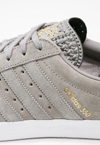 Grå mockaskor med tre perforerade ränder, en texturerad grå tåkappa och en gyllene "adidas 350"-logotyp, parat med en vit sula.