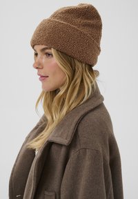 Beanie din fleece, pufos și de culoare maro, cu tiv pliat, asociat cu un palton din lână maro, cu guler cu vârf. Design texturat și călduros.