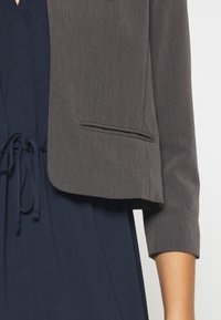 Harmaa lyhyt bleiseri, jossa on rakenteellinen muotoilu, vasemmalla puolella yksi tasku ja kolmanneksen pituiset hihat, sinisen mekon päällä.