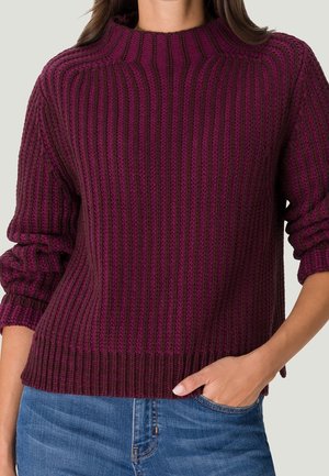 Pull en maille de couleur bordeaux foncé avec une texture côtelée, un large col rond et des poignets roulés, associé à un jean bleu.