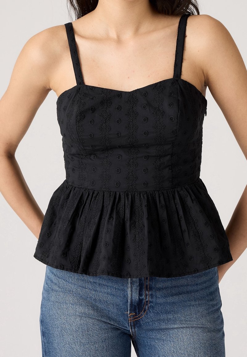 Femme portant un top péplum noir sans manches avec broderie florale associé à un jean denim bleu, debout devant un fond uni.