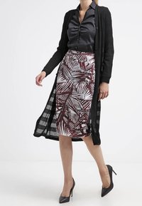 Femme portant un chemisier noir, un long cardigan noir, une jupe à motif arrivant aux genoux et des talons hauts noirs, marchant devant un fond blanc uni.