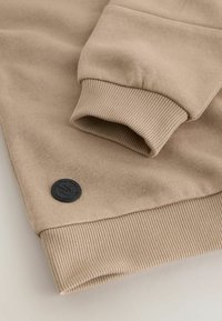 Beiger Sweatshirt-Stoff mit gerippten Bündchen und Saum, mit einem schwarzen runden Logopatch nahe dem Bündchen. Glatte Textur, lässiges Design.