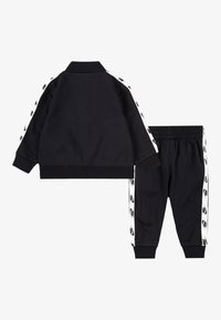 Fekete melegítőszett cipzáras dzsekivel és keskenyedő nadrággal. Mindkettő fehér oldalsávokkal díszített, fekete logós részletekkel.