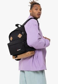 Mochila Eastpak en negro y marrón con acentos de diamante, correas ajustables y un compartimento principal con cremallera, presentada sobre un fondo blanco.