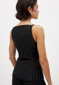 Ženska s kratkimi temnimi lasmi, oblečena v črn brezrokavni top s črtami in usklajene hlače, obrnjena stran od preprostega svetlega ozadja.