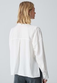 Weißes Langarmshirt mit vertikalen Streifen, leicht oversized Design und einer Knopfverschluss am hinteren Halsauschnitt. Glatte Textur, lässiger Stil.