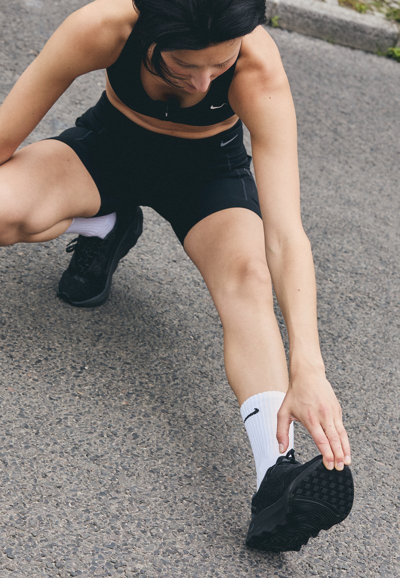 Vêtements de sport noirs, chaussettes blanches et chaussures noires. La personne s'étire en saisissant son pied, montrant une semelle de chaussure en caoutchouc texturé.