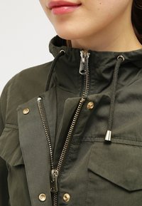 Kurtka oliwkowa z dwuścieżkowym zamkiem błyskawicznym, przednimi kieszeniami z zatrzaskami, kapturem ze ściągaczem oraz złotymi elementami ozdobnymi.
