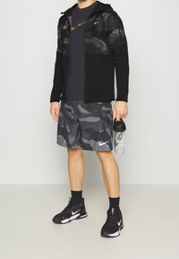 Chaqueta negra con cremallera y parte superior con patrón de camuflaje, pantalones cortos grises de camuflaje, calcetines negros y zapatillas, sosteniendo una botella de agua transparente.