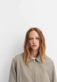 Lys grå zip-up jakke med krave, der har en sømdetaljering. Stoffet virker glat, og den har to frontlommer.