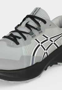 Chaussure de running ASICS gris clair avec lacets noirs, bandes logo blanches et noires, et une semelle noire épaisse avec une texture crantée.