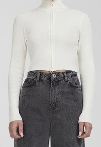 Vit ribbad zip-up långärmad topp som är kombinerad med högmidjade svarta jeans. Toppen har en figurnära silhuett och en hög krage.