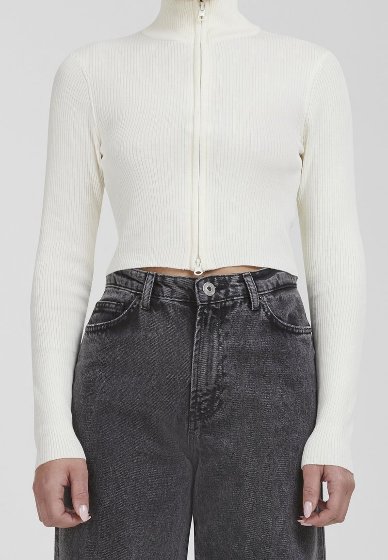 Vit ribbad zip-up långärmad topp som är kombinerad med högmidjade svarta jeans. Toppen har en figurnära silhuett och en hög krage.