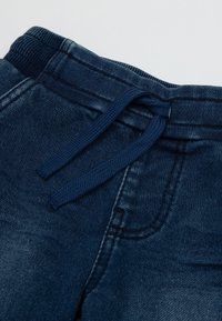 Pantaloni in denim blu scuro con un cinturino texturizzato, dotati di un cordoncino a fune e tasche, che mostrano un aspetto leggermente usurato.