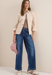 Veste en velours côtelé beige à boutons sur un haut beige, associée à un jean bleu marine à jams larges. Accessoirisée avec une casquette rose et des baskets blanches.