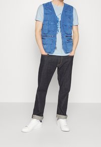 Modrý vícerozipový vest do světle modrého trička, v kombinaci s tmavými džíny a bílými teniskami. Obsahuje zipy a různé styly kapes.