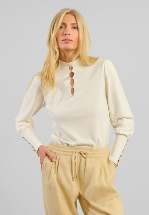 Pullover crema con collo alto e dettagli a chiave. Presenta maniche a sbuffo e bottoni decorativi sui polsini. Pantaloni beige a destra.