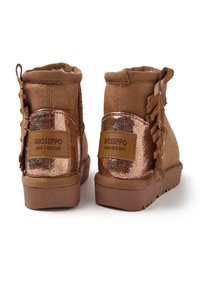 Botines de ante marrón con acentos metálicos en bronce, detalles de borlas y un panel trasero texturizado. Cuentan con una suela de goma.