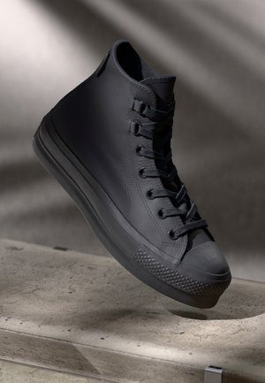 Black high-top sneaker with textured sole, laces tied, suspended above a concrete ledge in soft shadowed lighting.