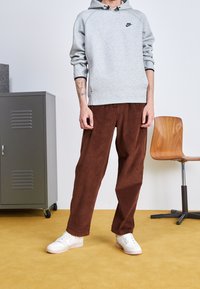 Persona vestida con sudadera gris de Nike, pantalones de pana marrón y zapatillas blancas de pie sobre un suelo amarillo cerca de un armario gris y una silla de madera.
