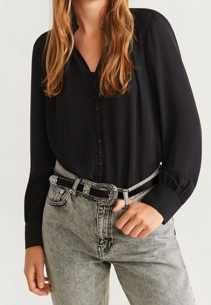 Blusa nera con scollatura a V, chiusura con bottoni e maniche lunghe. Abbinata a jeans grigi sbiaditi e una cintura nera con dettagli metallici.