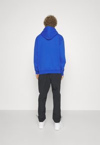 Modrá mikina s kapucí a kapsou jako klokan, spárovaná s černými cargo kalhotami. Bílé tenisky dotvářejí outfit, pohled ze zad.