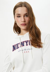 Witte geribde hoodie met "NEWYORK" in rode en blauwe letters, voorzien van een capuchon met trekkoord en de opdruk "Living the dream 1995."