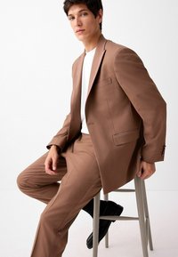 Costume marron ajusté avec une veste structurée et un pantalon à plat, associé à une chemise blanche. Texture lisse et design classique, assis sur un tabouret.